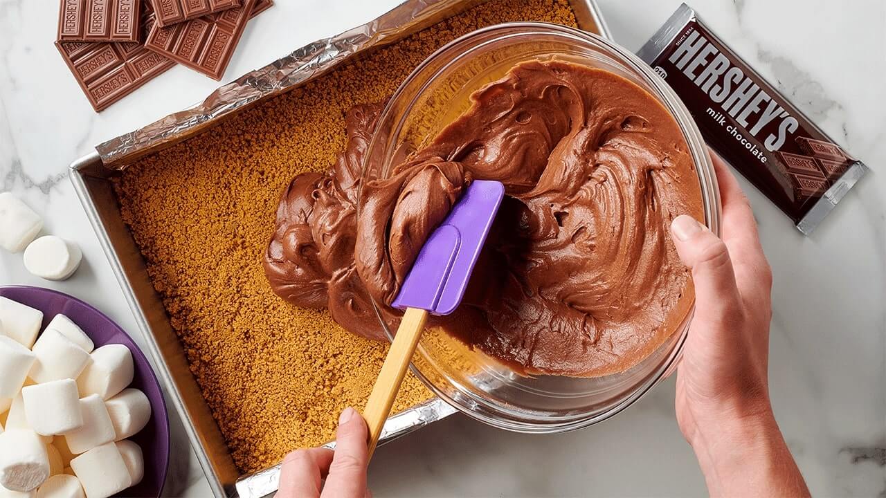 person spreading brownie mixture on top of a graham cracker crust